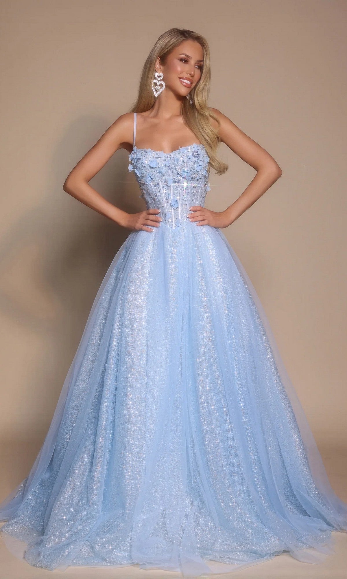A woman in the blue Portia and Scarlett PS26146 Glitter Prom Ball Gown with spaghetti straps, a boned-corset bodice, glitter tulle, and floral details poses against a plain beige background wearing large heart-shaped earrings.